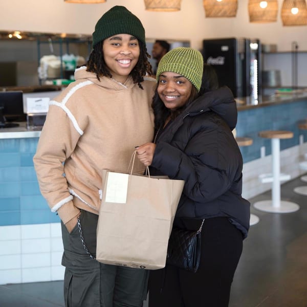 Two smiling women in Yumbii restaurant, one holding a takeout bag, ready for Mexican food.