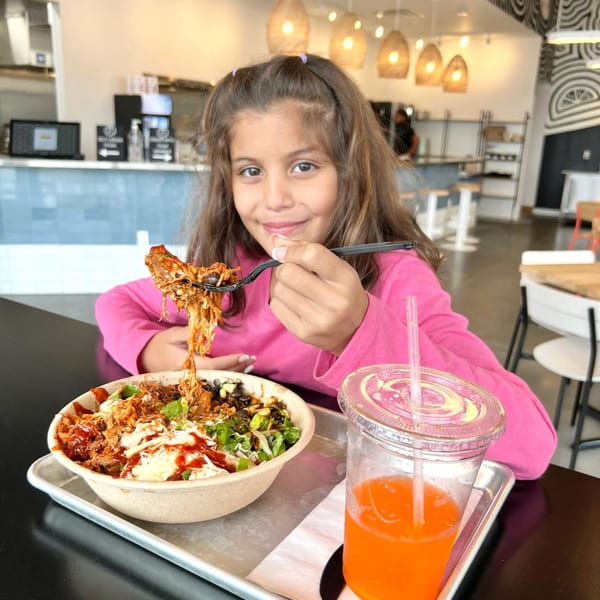 A young girl in a pink shirt smiles while eating a Yumbii Mexican food bowl.