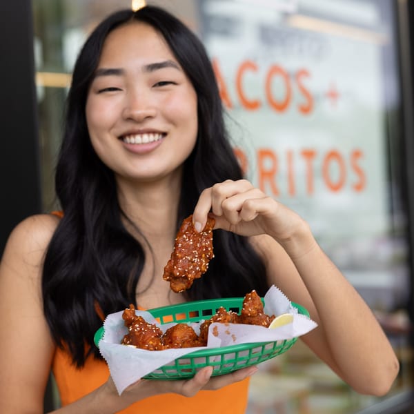 Smiling woman holding a green basket of Yumbii's sesame-crusted chicken wings, with a taco sign in background.