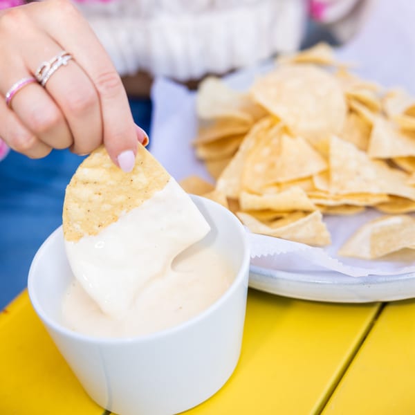 A hand dips a tortilla chip into a white bowl of Yumbii's creamy queso, with more chips in the background.