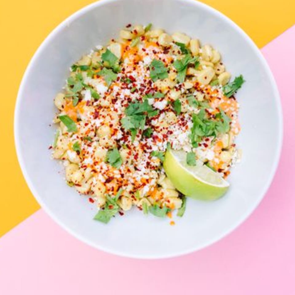 Yumbii's Mexican street corn in a white bowl, topped with cheese, chili flakes, cilantro, and lime.