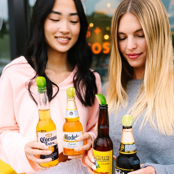 Two women holding various Mexican beers with lime wedges, smiling in front of a restaurant.