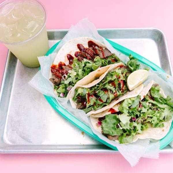 Three Yumbii tacos with meat, lettuce, and red sauce, served with a lime wedge and drink.